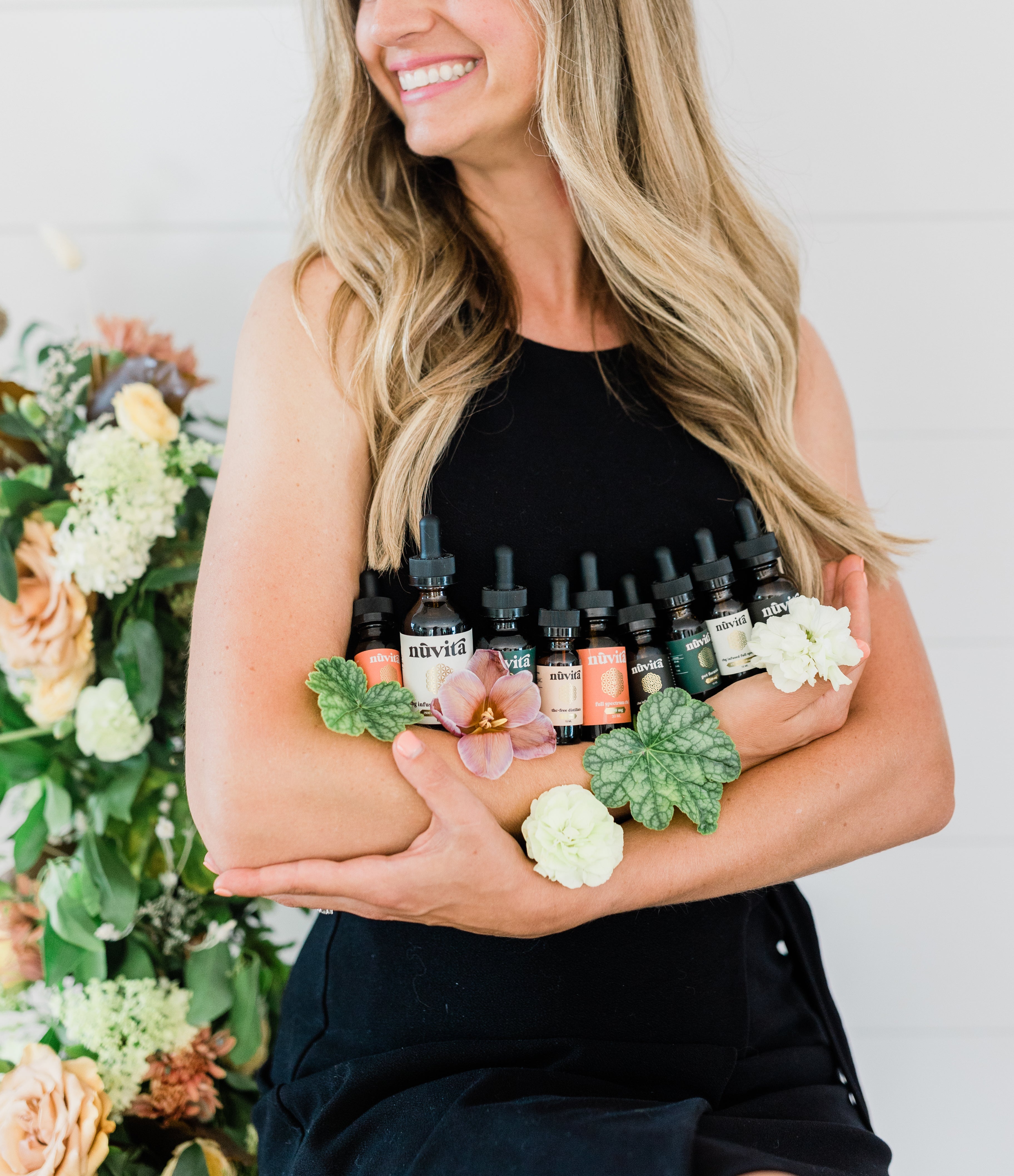 Woman cradling Nuvita's array of tinctures standing a room with flowers decorating the background